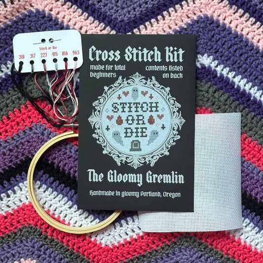 A cross stitch kit laid out on a chevron stripe crochet blanket. The kit packaging reads 'The Gloomy Gremlin' and 'Cross Stitch Kit'. Also displayed is a white thread chart with colored threads, a wooden embroidery hoop, and a white embroidery fabric.