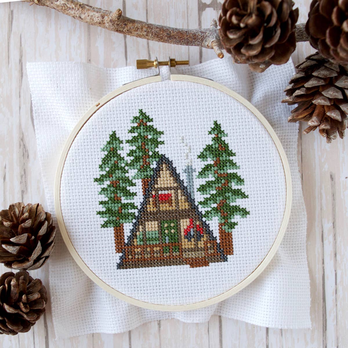 A finished project on a wooden design background with a branch and several pine cones surrounding the embroidery hoop.