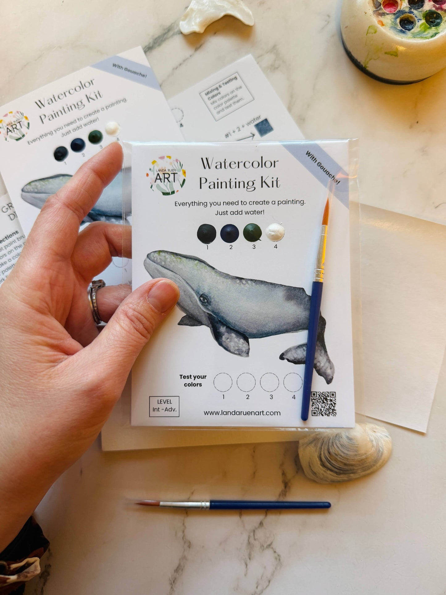 Watercolor painting kit with whale illustration held in hand with a marble surface in the background.