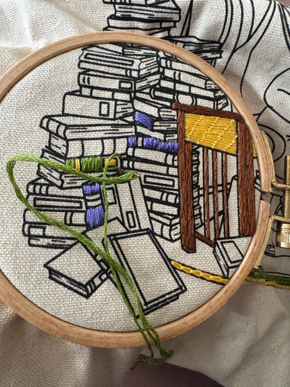 Close up view of an embroidery hoop with a detailed embroidery of books and a side table.