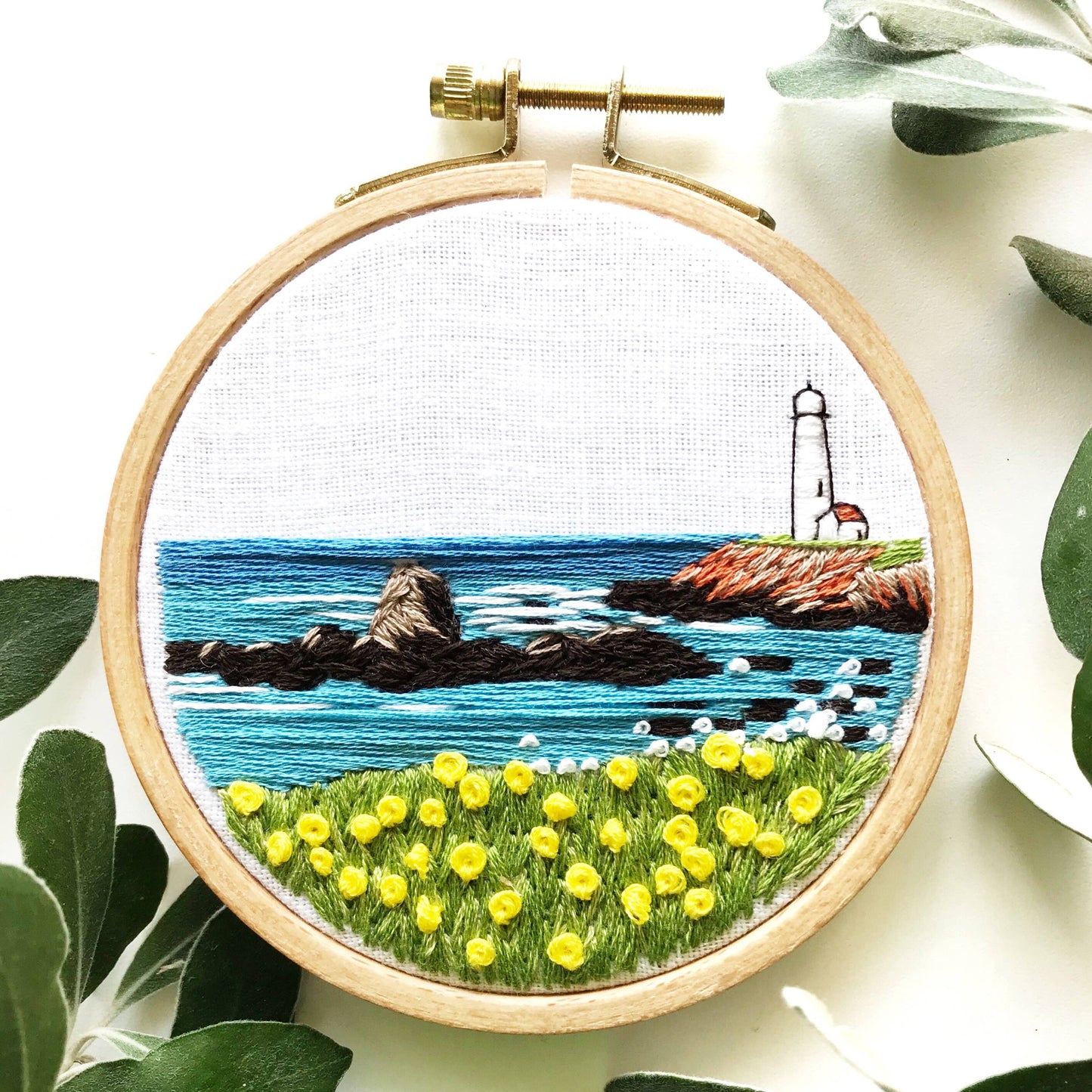 Embroidery hoop with a scenic landscape design featuring a lighthouse, ocean, and grass with yellow flowers.