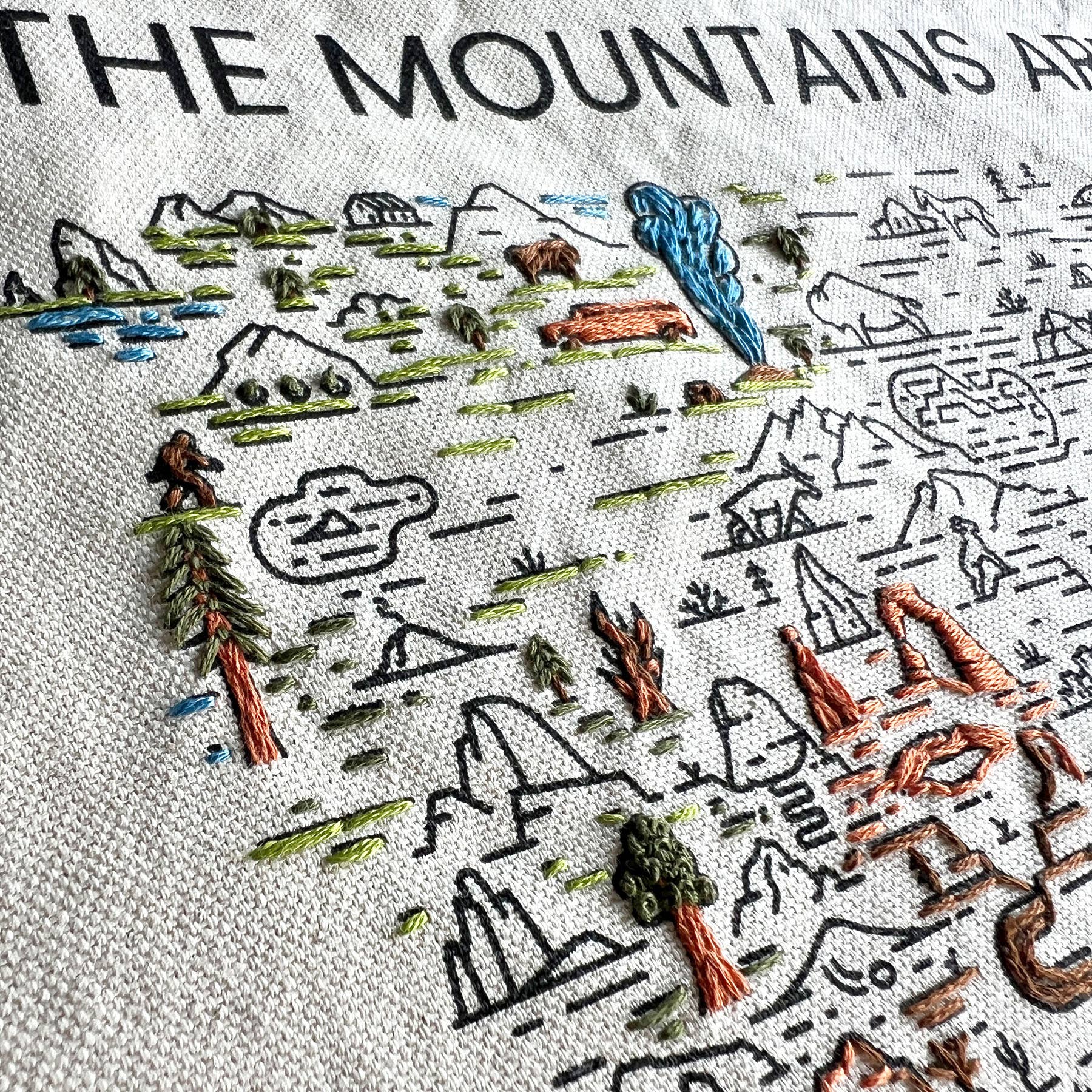 A close up view of a partially embroidered example of the mountains and nature design on fabric with text 'The Mountains Are Calling'.
