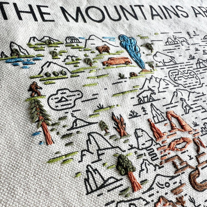 A close up view of a partially embroidered example of the mountains and nature design on fabric with text 'The Mountains Are Calling'.