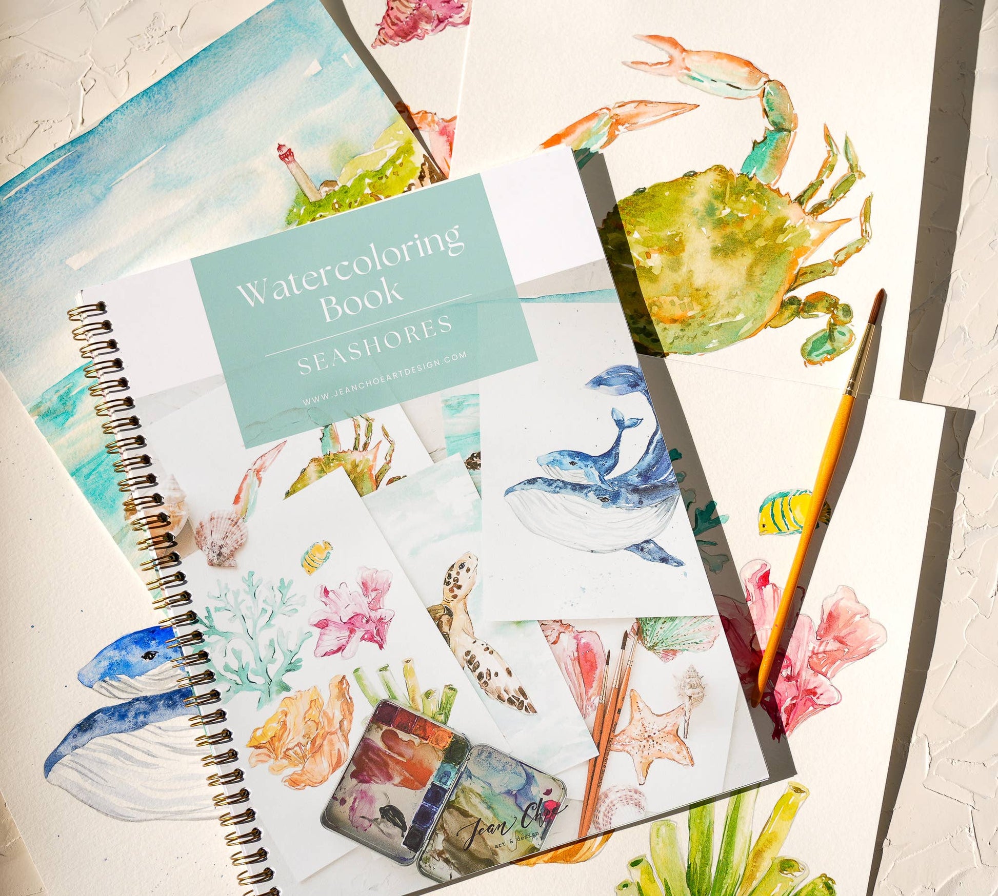 Watercolor workbook on top of a collection of completed watercolor paintings of the included designs. On a textured surface.