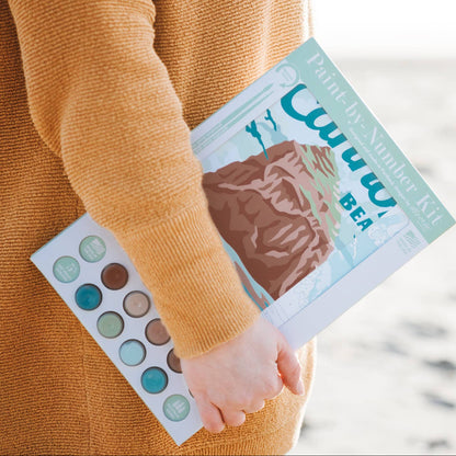 Person holding the paint-by-number kit on a blurred beach background