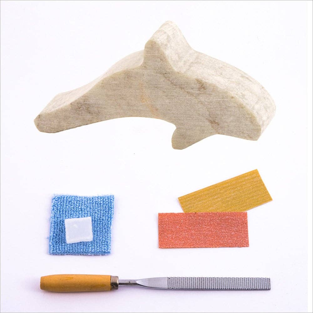 The rough cut Soapstone orca shape, with two grades of sandpaper, a carving file, polishing wax and a blue buffing cloth in front of a white background.