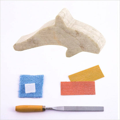 The rough cut Soapstone orca shape, with two grades of sandpaper, a carving file, polishing wax and a blue buffing cloth in front of a white background.
