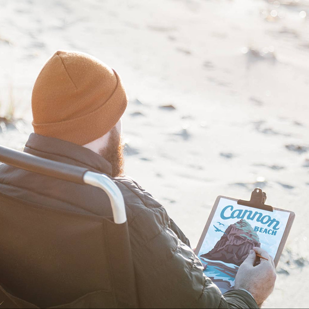 Person sitting on a chair on a sandy beach, holding a paintbrush painting cannon beach. 