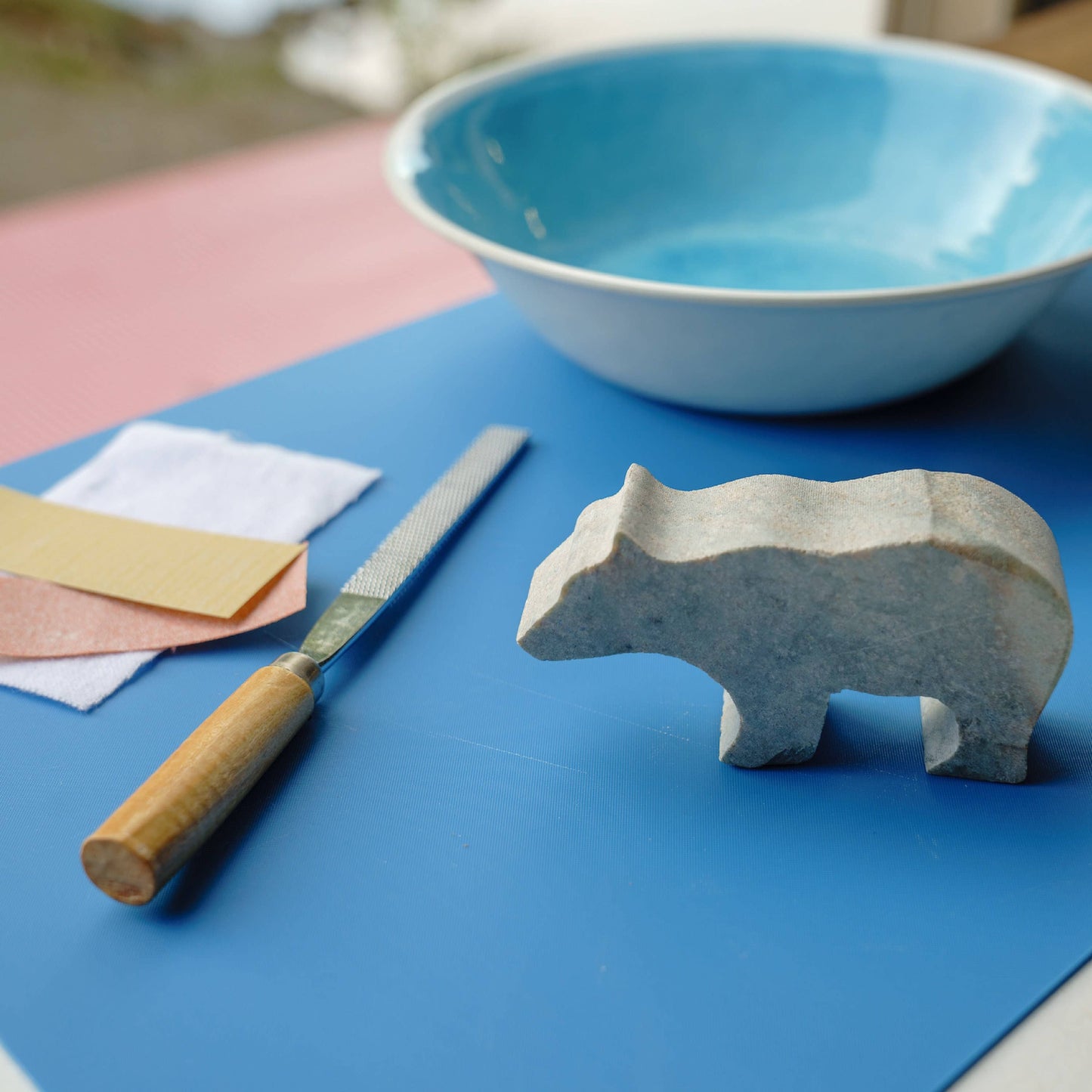 The rough cut starting shape of the bear figure, with sandpaper, a cloth, carving file, and light blue bowl on a darker blue surface. 