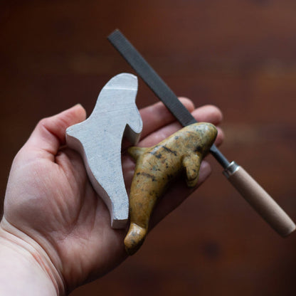A hand holding a stone carving file and two stone carvings, one in the original rough cut shape and one in the finished state after being shaped and polished. 