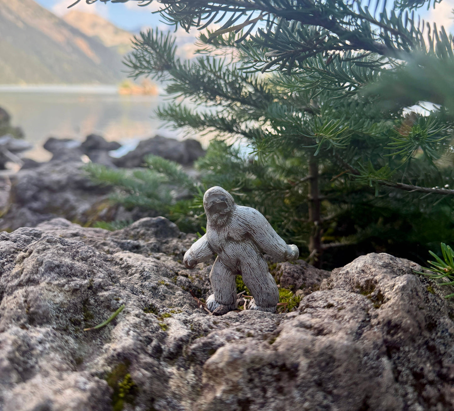 A finished Bigfoot soapstone carving in a nature setting. Bigfoot appears on top of hills of stone, and surrounded by tree branches. 