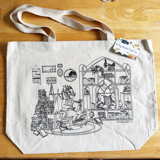 A canvas tote bag laid flat on a wood table. The bag has a line drawing of a dragon reading while sitting in a home library surrounded by books. 
