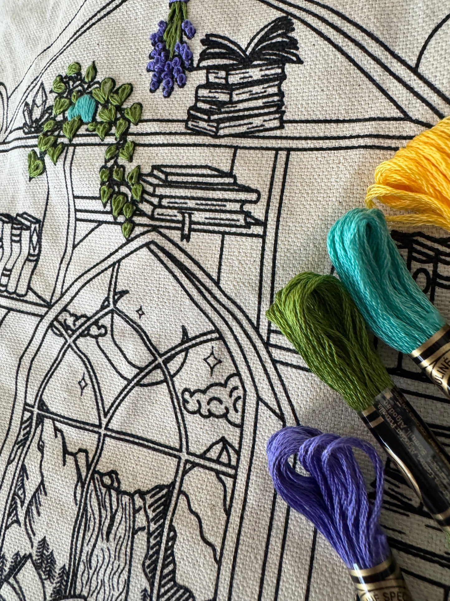 Close up view of a partially completed embroidery design with black outlined design, of a shelf laden with books, plants and flowers on a canvas background. Laid across the design are four skeins of embroidery thread in yellow, robins egg blue, green, and lavender.