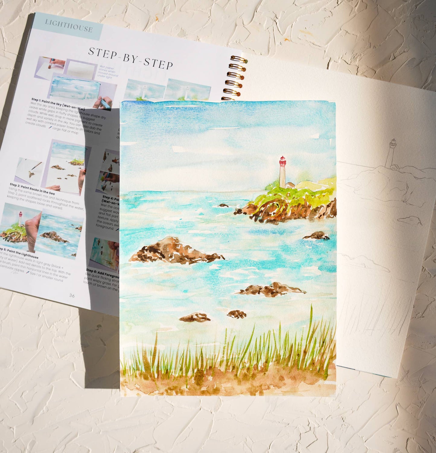 A lighthouse watercolor painting next to the workbook open on step by step instructions. The background is a neutral textured surface