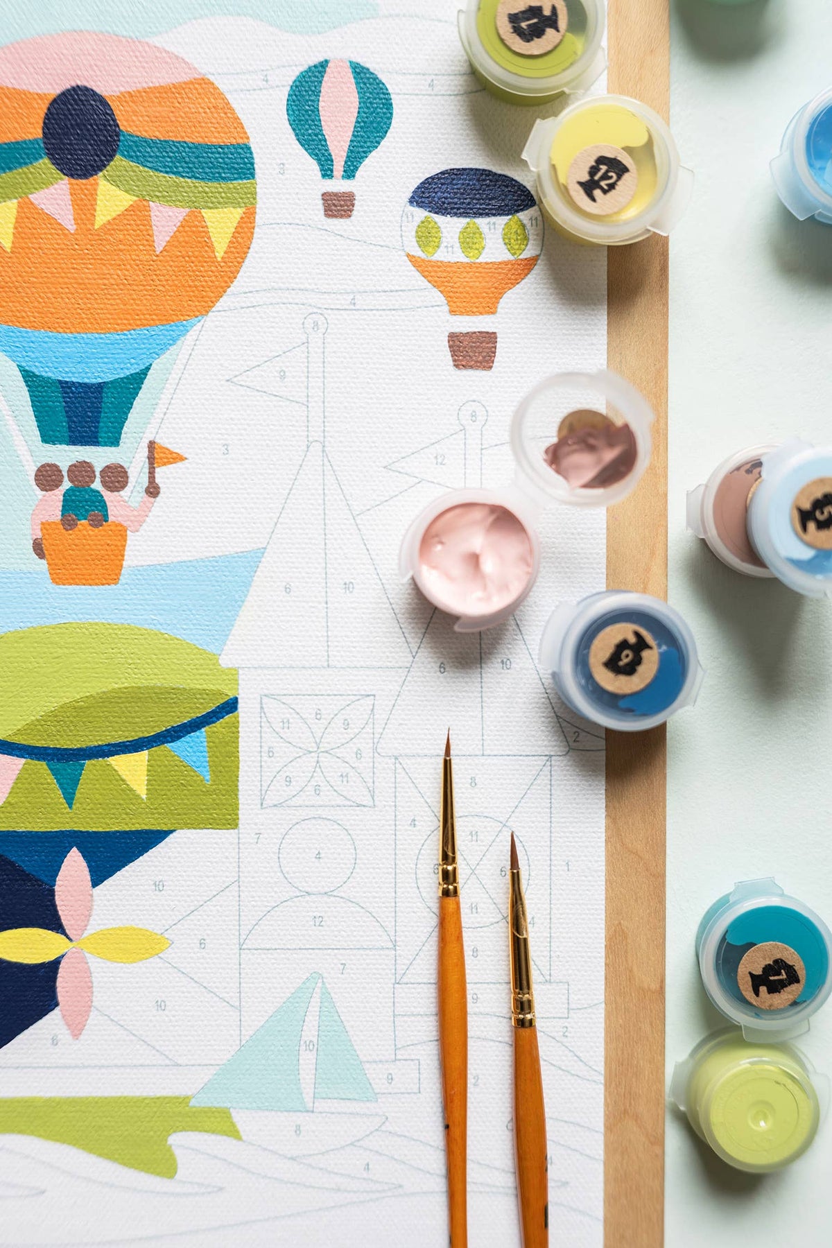 Paint by numbers painting with hot air balloons and castle design, surrounded by paint containers and brushes on a light background.