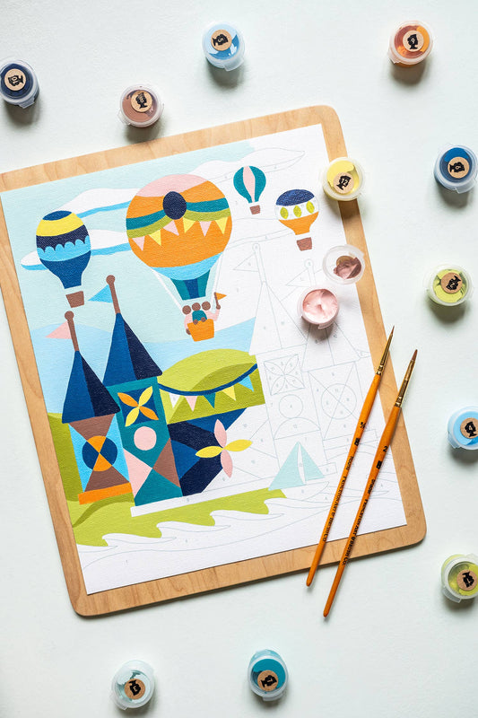 Paint by numbers painting with hot air balloons and castle design, surrounded by paint containers and brushes on a light background.