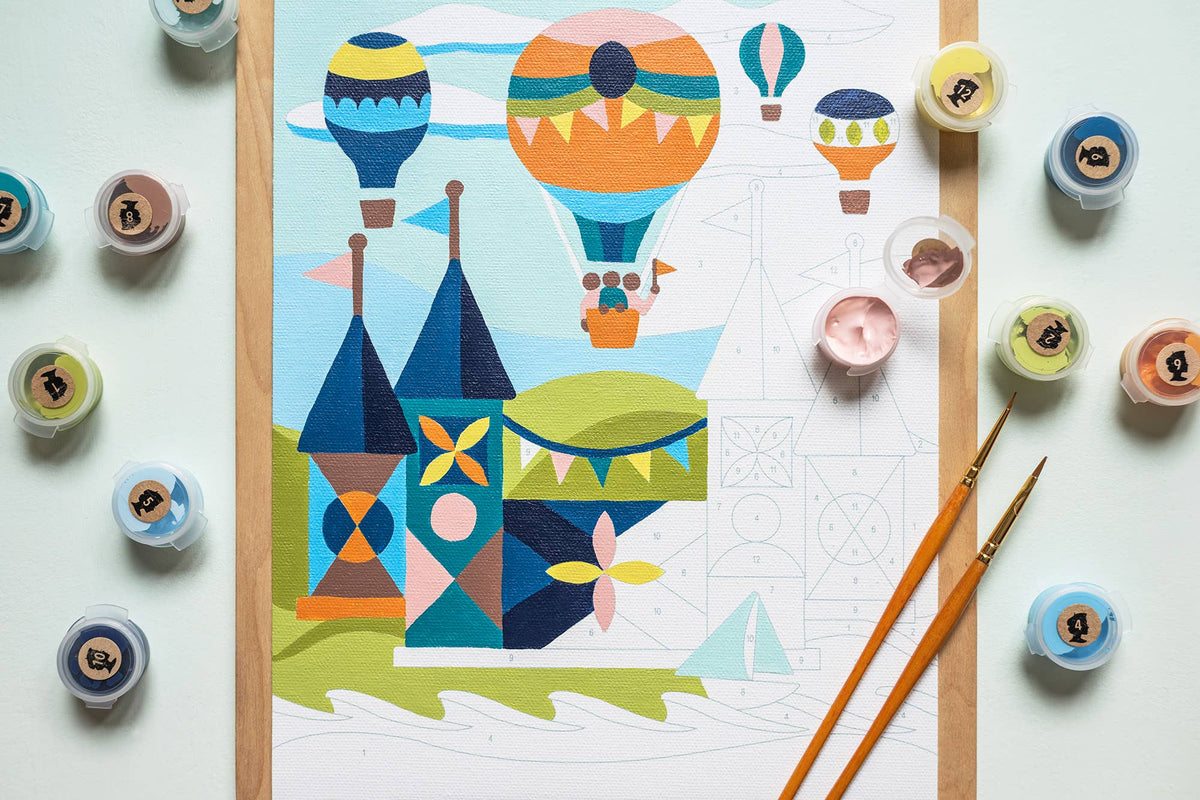 Paint by numbers painting with hot air balloons and castle design, surrounded by paint containers and brushes on a light background.