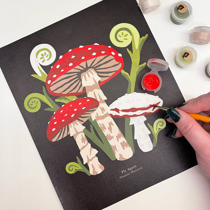 Person painting a detailed illustration of mushrooms on a black canvas with art supplies around.