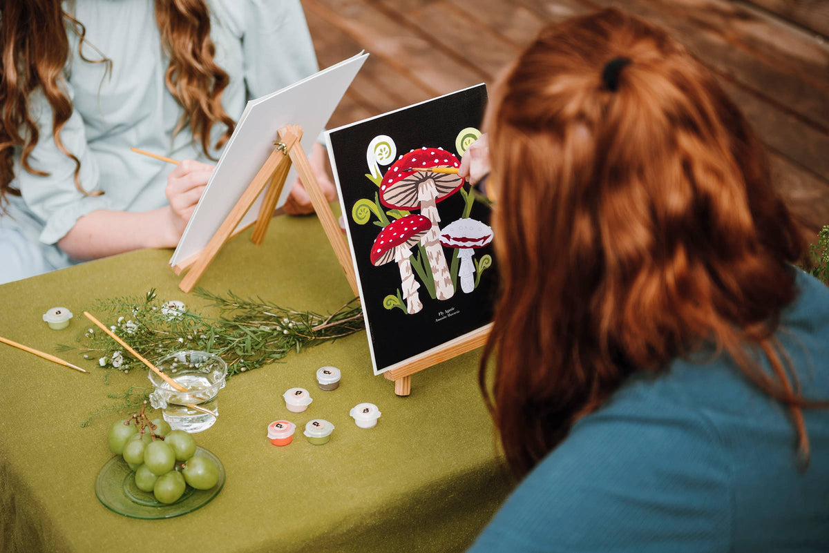 Two people sitting at a table with art supplies and a painting of mushrooms.