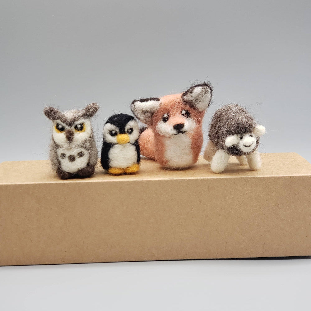 Four small animal figurines (owl, penguin, fox, sheep) on a cardboard box against a gray background.