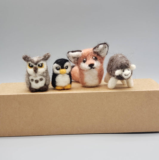 Four small animal figurines (owl, penguin, fox, sheep) on a cardboard box against a gray background.