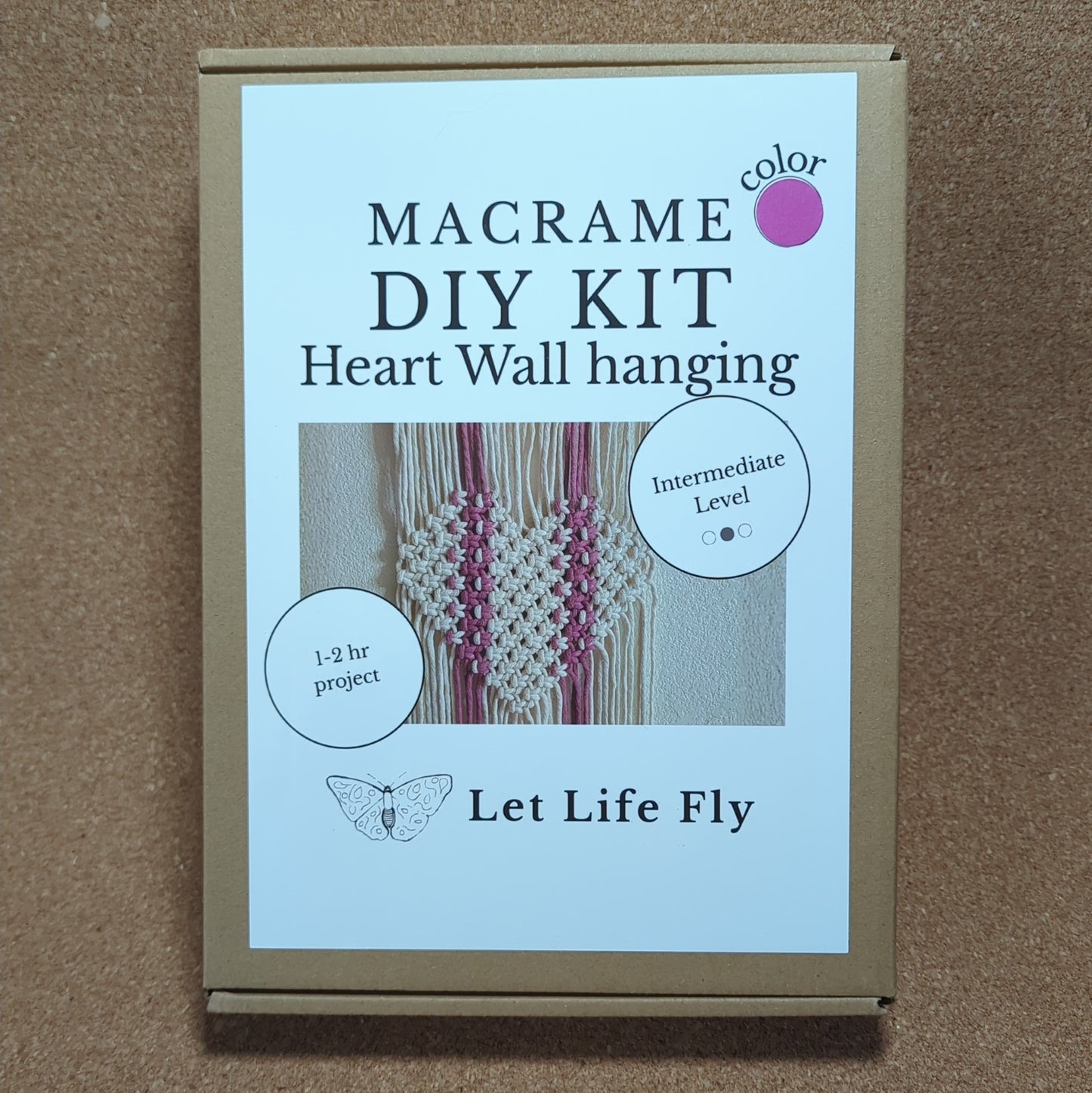 Macrame DIY kit packaging with text and a small image of a heart wall hanging on a cork background