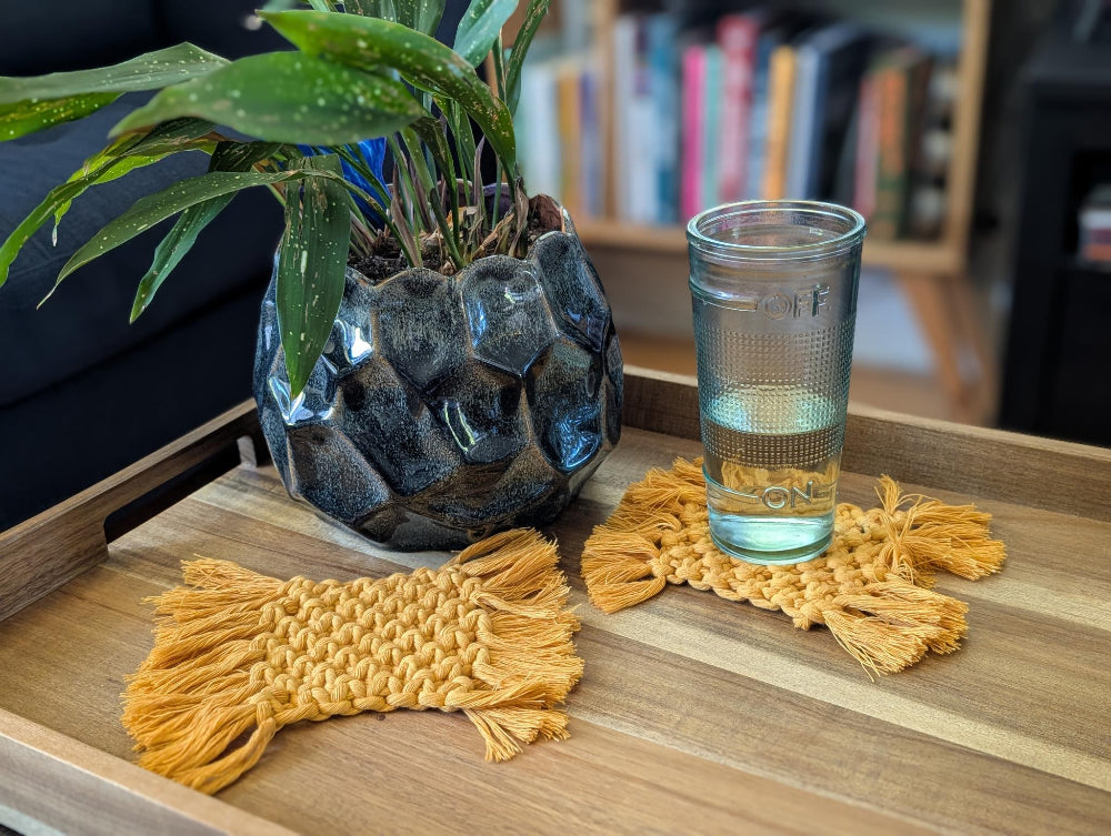 Glass of water on a wooden tray with decorative macrame coasters and a plant in the background.