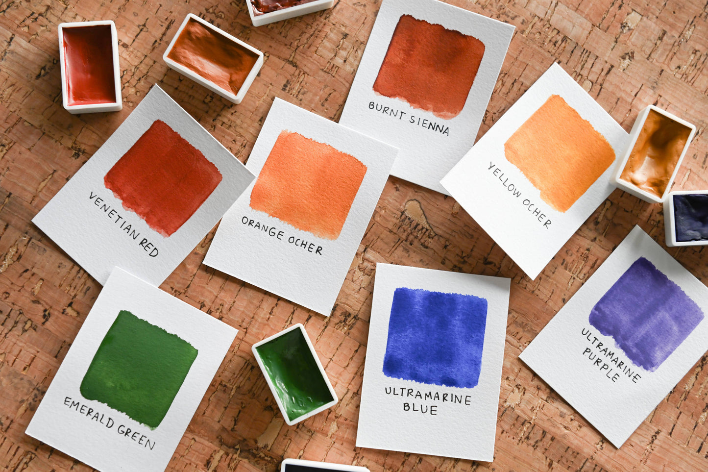 Color swatches on watercolor paper with the color names next to small pans of watercolor paint on a cork surface