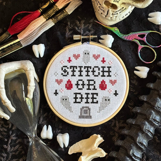 Cross-stitch hoop with 'Stitch or Die' text surrounded by embroidery tools on a dark background