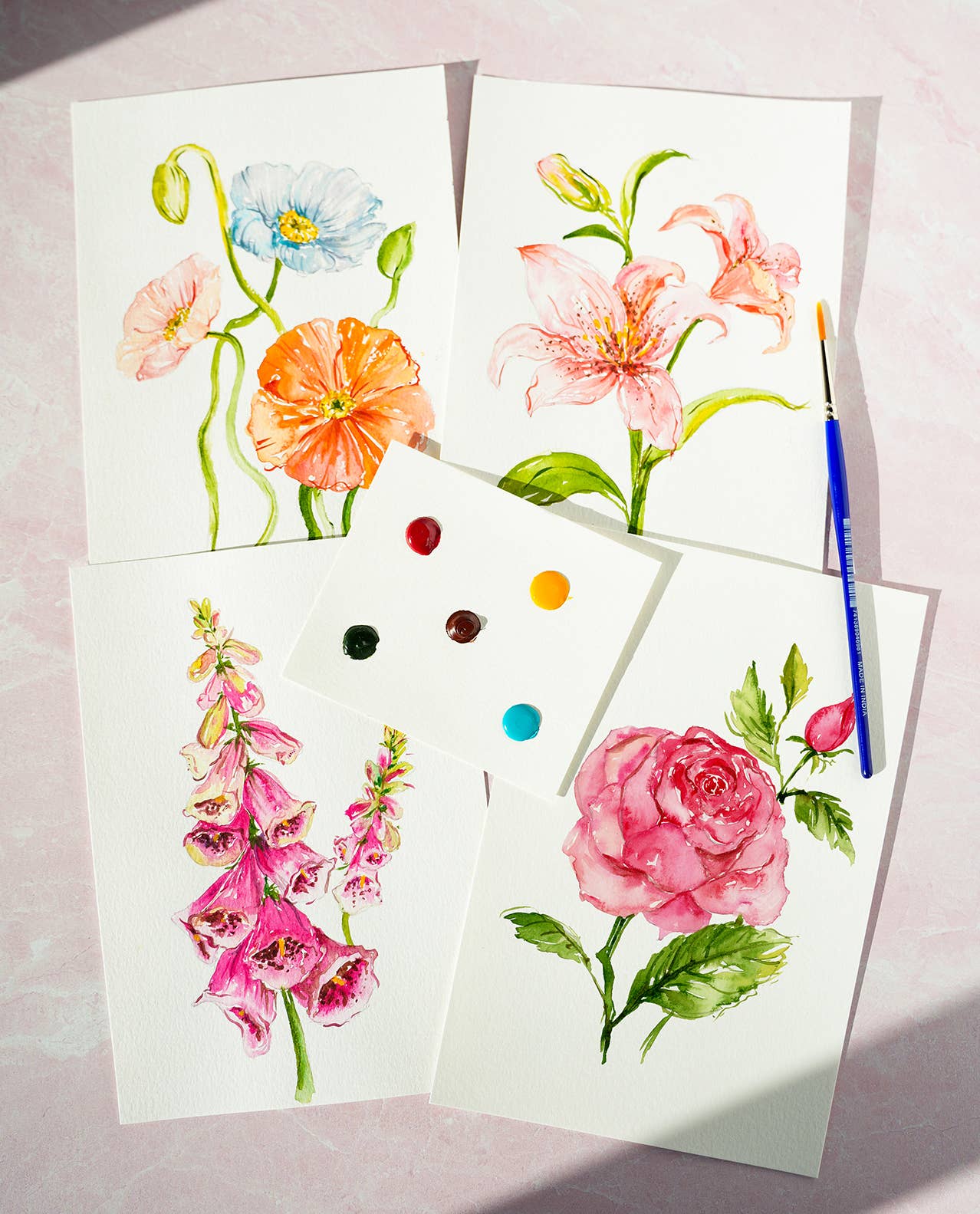 Finished watercolor flower paintings and a paintbrush on a light pink background