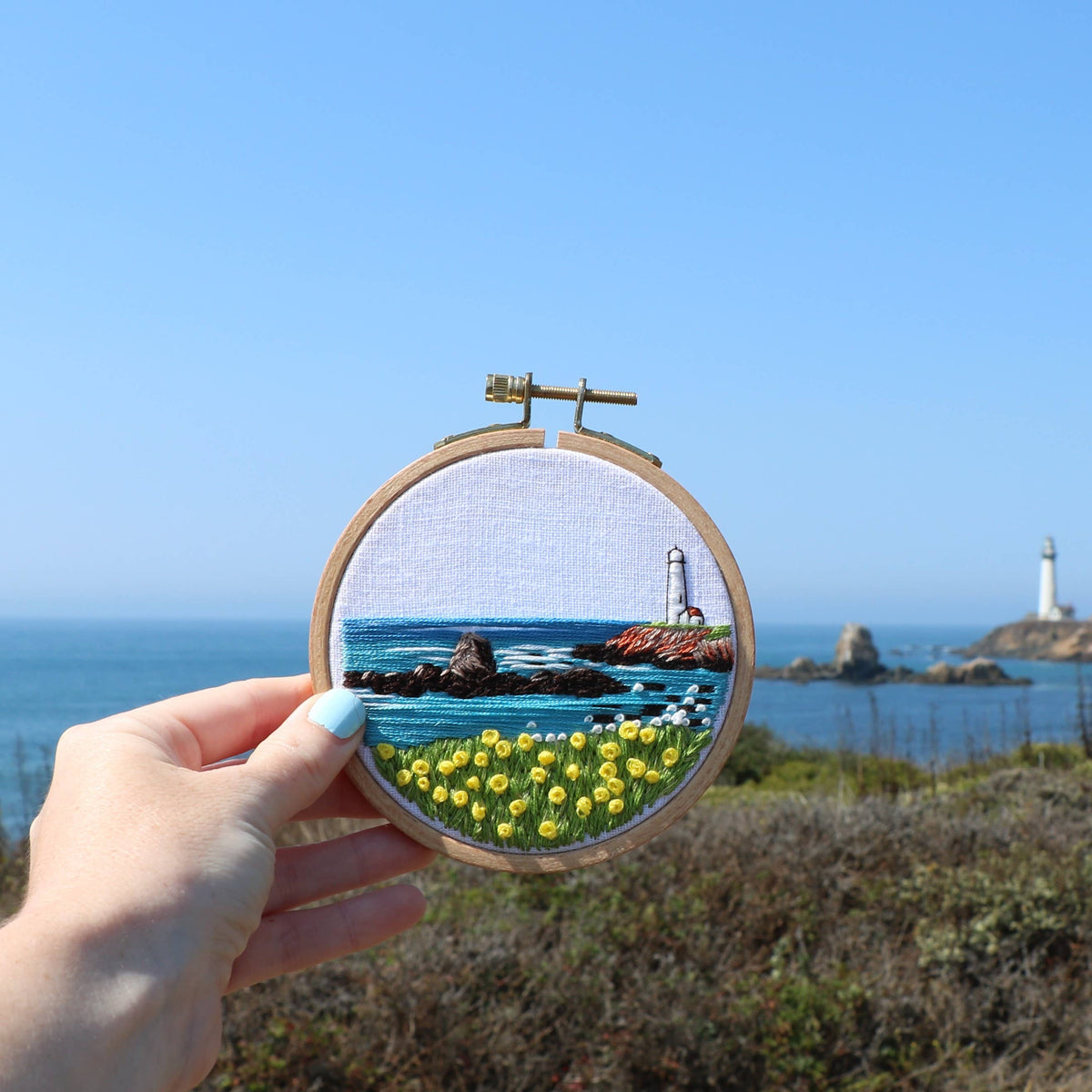 Embroidery hoop with a scenic landscape of a lighthouse and ocean held up so that in the background is the real-life coastal background it was designed on. 