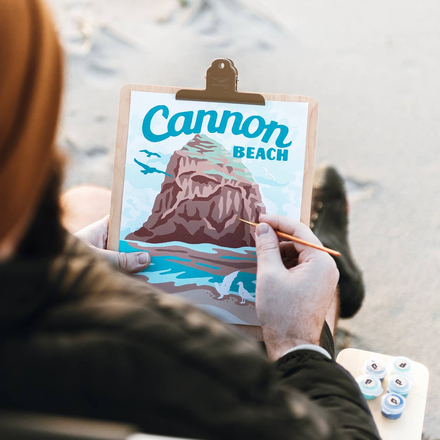 A man at the beach, holding a paintbrush and a finished painting of Cannon Beach