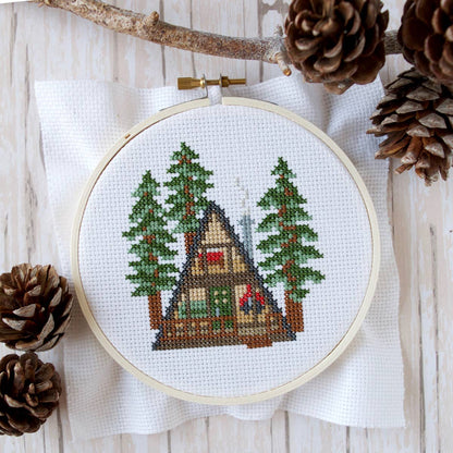 A finished project on a wooden design background with a branch and several pine cones surrounding the embroidery hoop. 