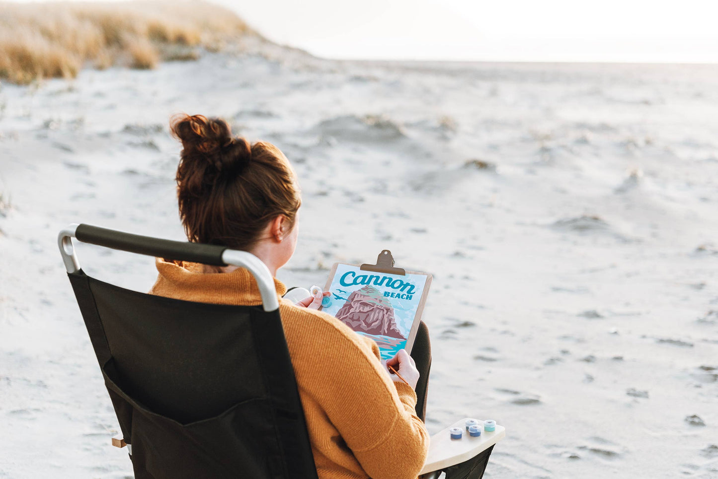 Person sitting in a chair at the beach holding a paintbrush painting a picture of the notable rock at Cannon Beach