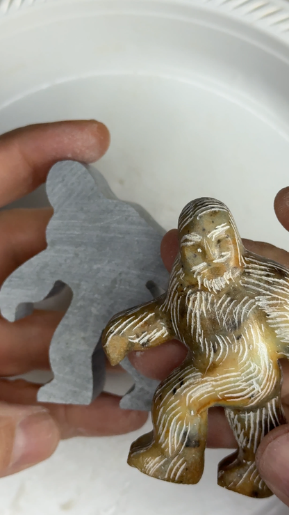 A rough cut starting shape of Bigfoot  next to a finished Bigfoot soapstone carving. Held in two hands with the finished slightly in-front of the rough cut. 