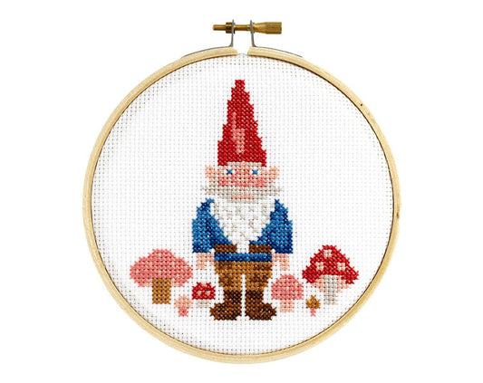 The finished project displayed in the embroidery hoop. The design shows a rosy cheeked bearded gnome wearing a red cone hat, and a blue top over brown pants and darker brown boots. Mushrooms on either side have red and pink tops with lighter spots. 