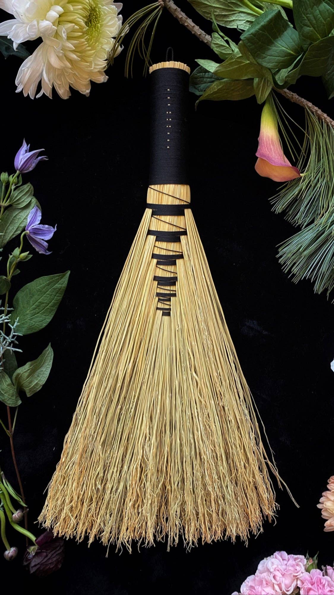 Traditional hand broom with black handle and broomcorn bristles against a dark background with flowers.