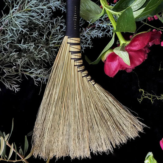 Broom with decorative handle on a dark background with greenery and pink flowers