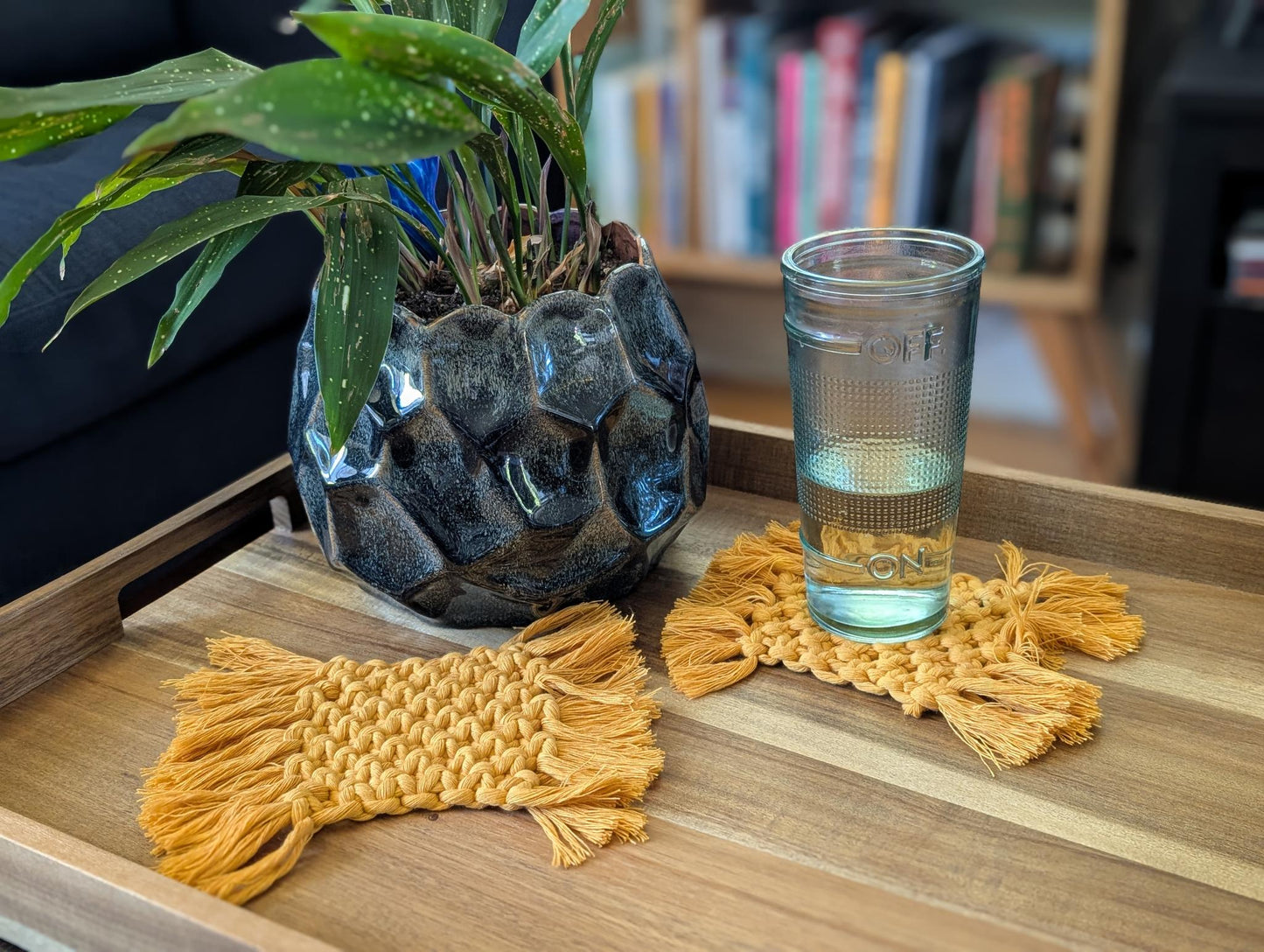 Glass of water on a wooden tray with decorative macrame coasters and a plant in the background.