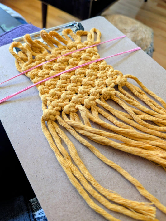 Yellow macrame project in progress on a wooden clipboard held inplace with pink rubberbands