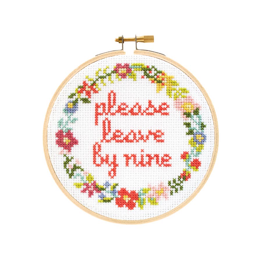 A cross stitch project in a wooden hoop. The text 'please leave by nine' is circled by a floral design. White background. 